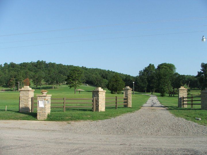 Stone entrance gates with a gravel road leading to a green space with trees and a light blue sky.