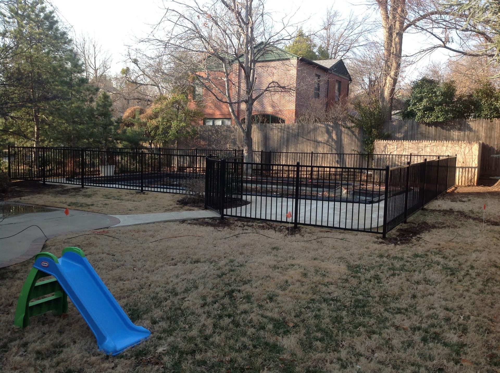 Backyard with black fence, blue slide, and brown grass. Brick house and trees in background.