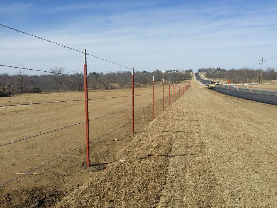 Barbed wire fence with red metal posts along a dirt path on a sunny day.