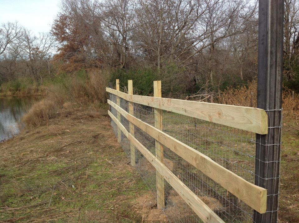Wooden fence with wire mesh near a body of water, trees in the background.