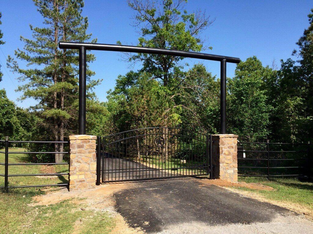 Black metal gate and archway at a driveway entrance, flanked by stone pillars and greenery.
