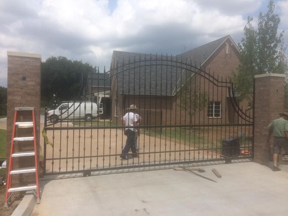 Iron gate spanning a concrete driveway, two brick pillars, two men, and a house in the background.