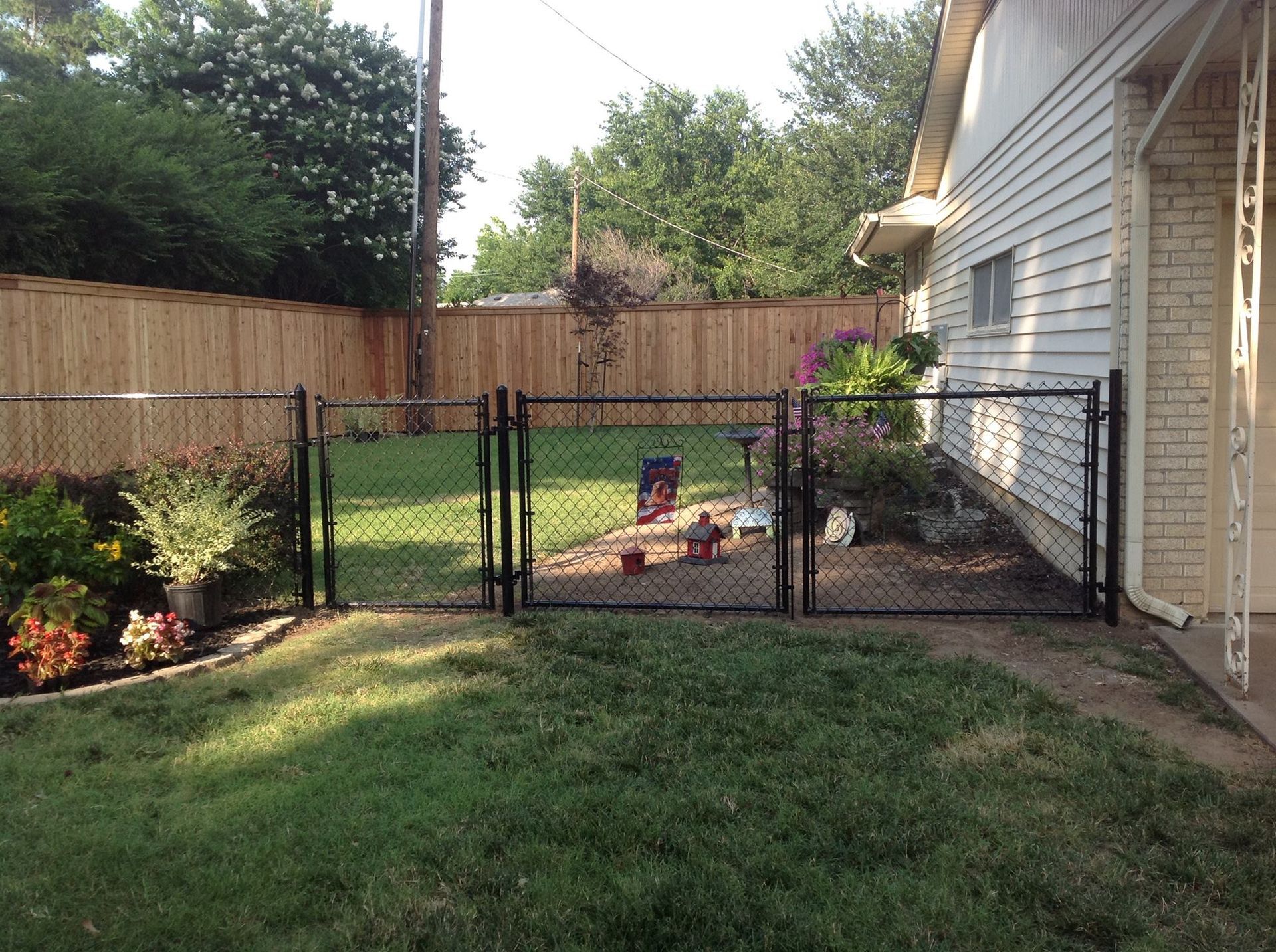 Black chain-link gates leading to a backyard garden, flanked by a wooden fence and house.
