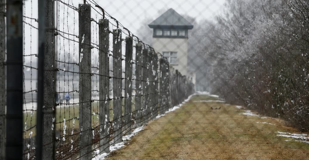 Barbed wire fence with guard tower in background, likely a concentration camp site.