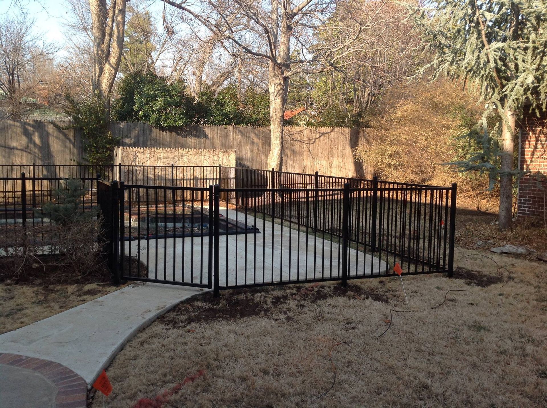Black metal fence surrounds a concrete patio in a backyard with trees and a wooden fence in the background.
