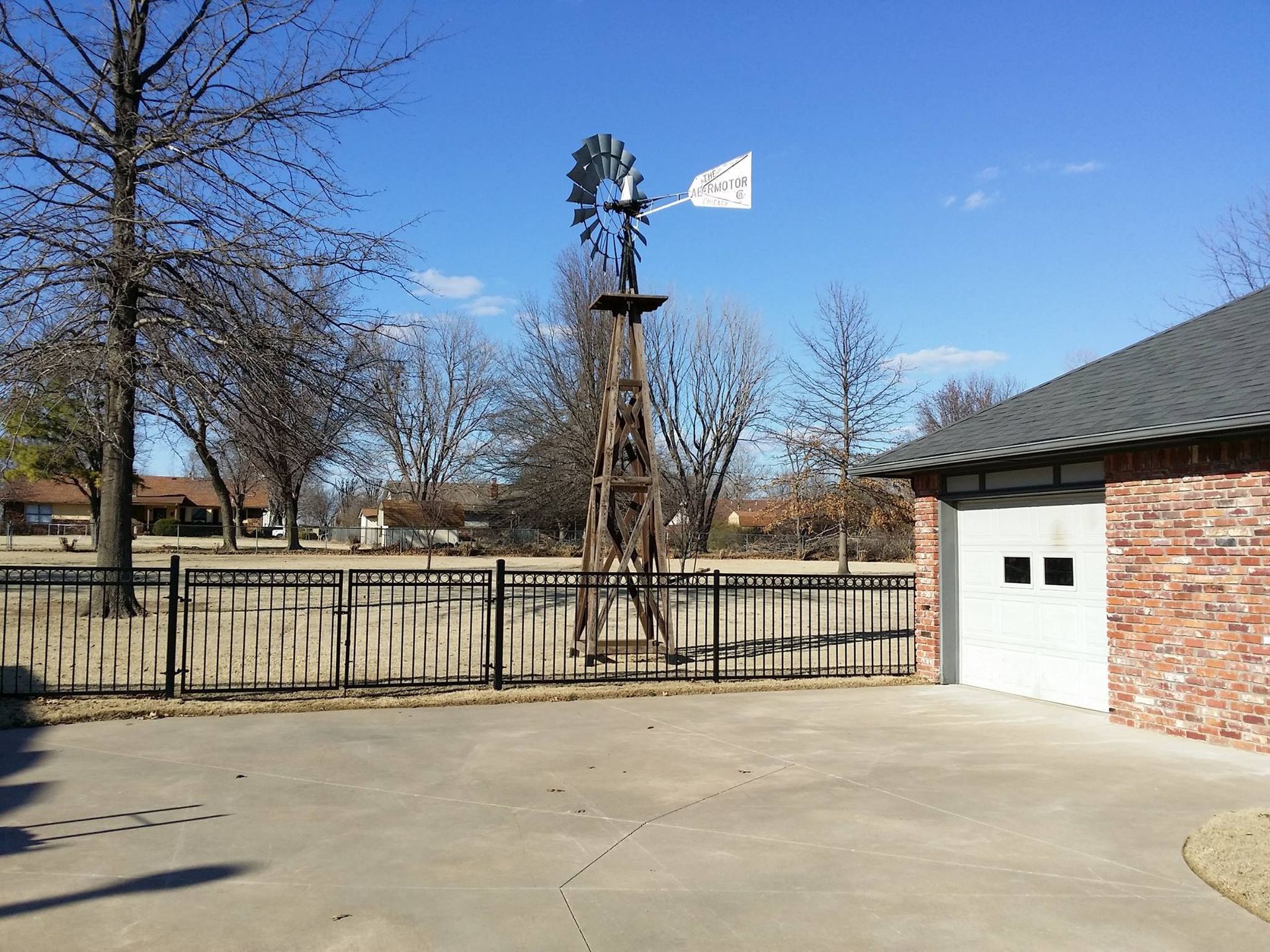 Windmill on a wooden tower next to a brick building and a black fence under a blue sky.