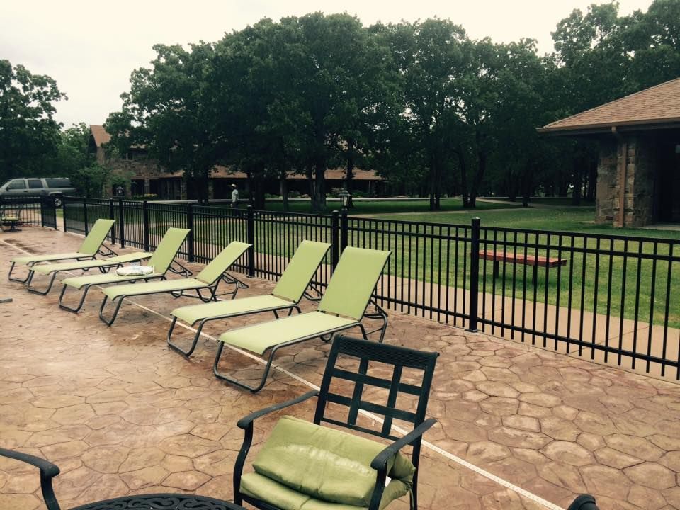 Lounge chairs near a pool with a black fence, trees, and a building in the background.