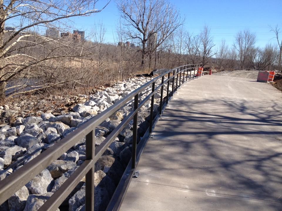 A paved pathway with a metal railing along a rocky embankment. Trees and buildings in the background.