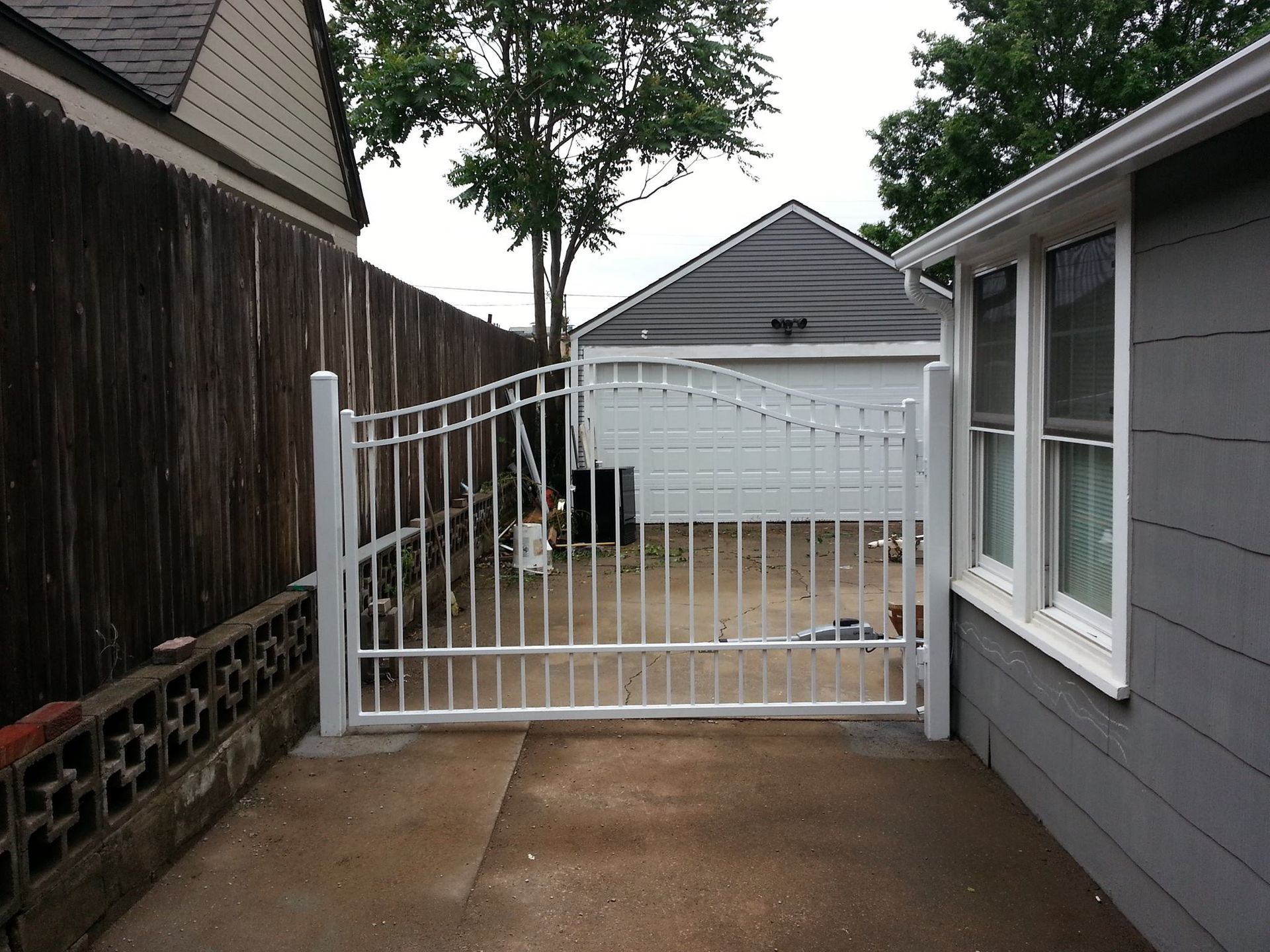 White metal gate in a driveway, leading to a garage. Gray siding and a wooden fence are visible.