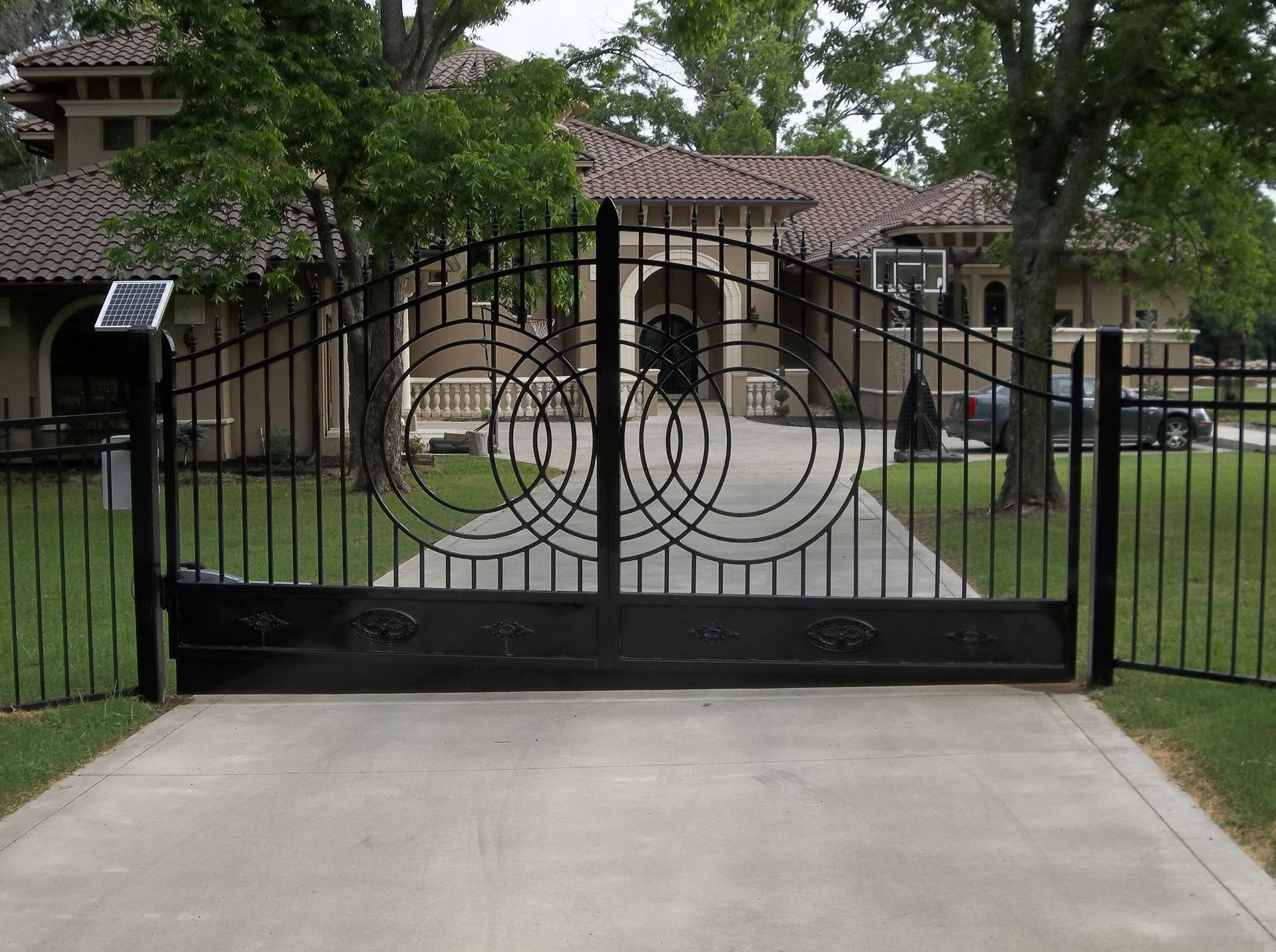 Black wrought iron gate with circular designs, on a driveway leading to a large house.