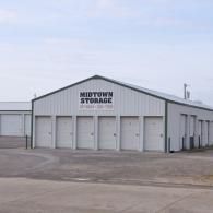 A white, metal-sided Midtown Storage building with multiple roll-up garage doors under a cloudy, light blue sky.