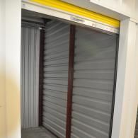 A storage unit with a closed grey metal roll-up door framed in dark brown trim and a yellow header.