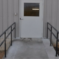 A concrete wheelchair ramp with black metal handrails leading to a white door on a gray metal-sided building.