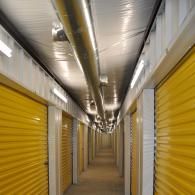 A long, narrow indoor storage facility corridor lined with closed yellow metal roll-up doors under bright overhead lights.