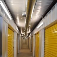 A corridor in a self-storage facility featuring yellow roll-up doors on both sides, bright overhead lights, and a duct.