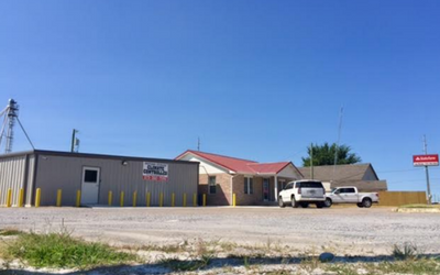 A gravel lot in front of a tan metal warehouse, a brick house with a red roof, and a sign under a clear blue sky.