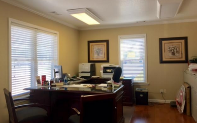 A wood-toned office with a desk, chairs, computer equipment, and two framed prints on the beige walls near two windows.