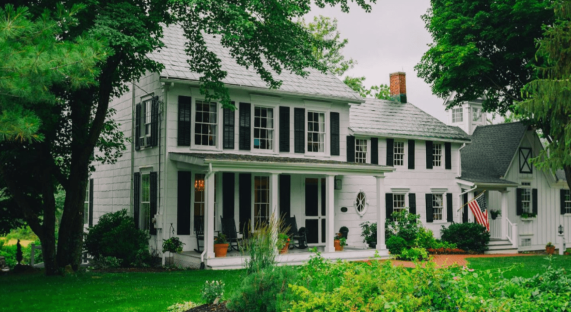 A large white house with a large porch is surrounded by trees.