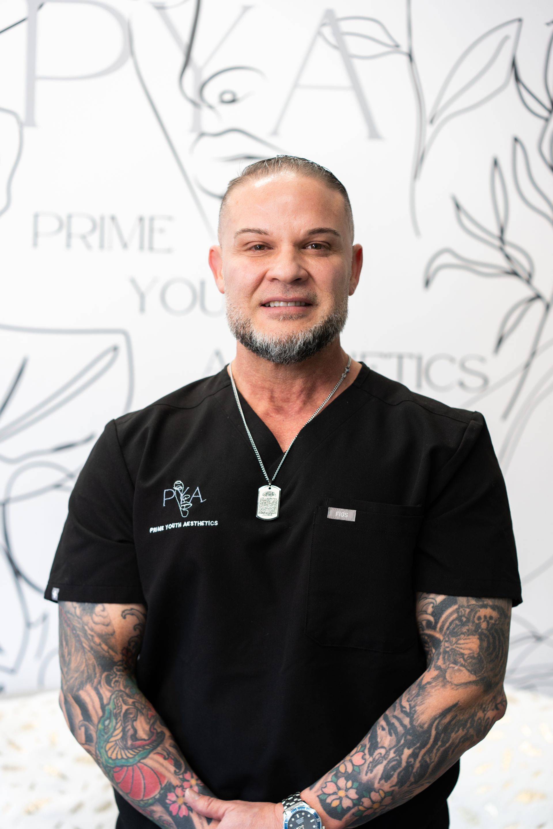 A man with a beard and tattoos is wearing a white shirt and a necklace.