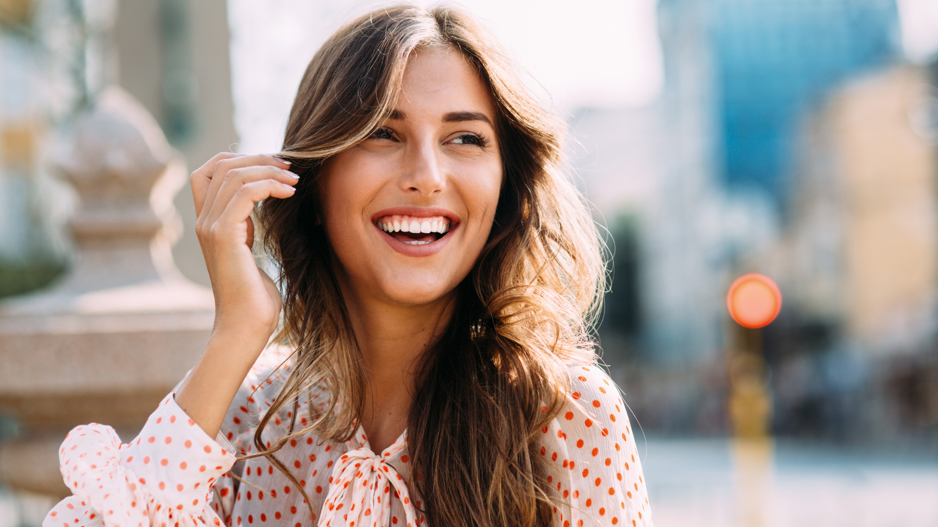 Woman with long brown hair smiles outdoors, touching her hair. Buildings and a traffic light are visible.