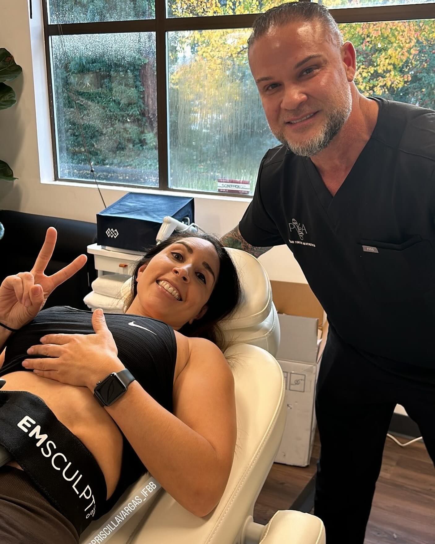 A man is standing next to a woman in a chair giving a peace sign.