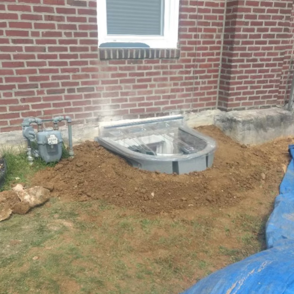 A window well is being installed on the side of a brick house.