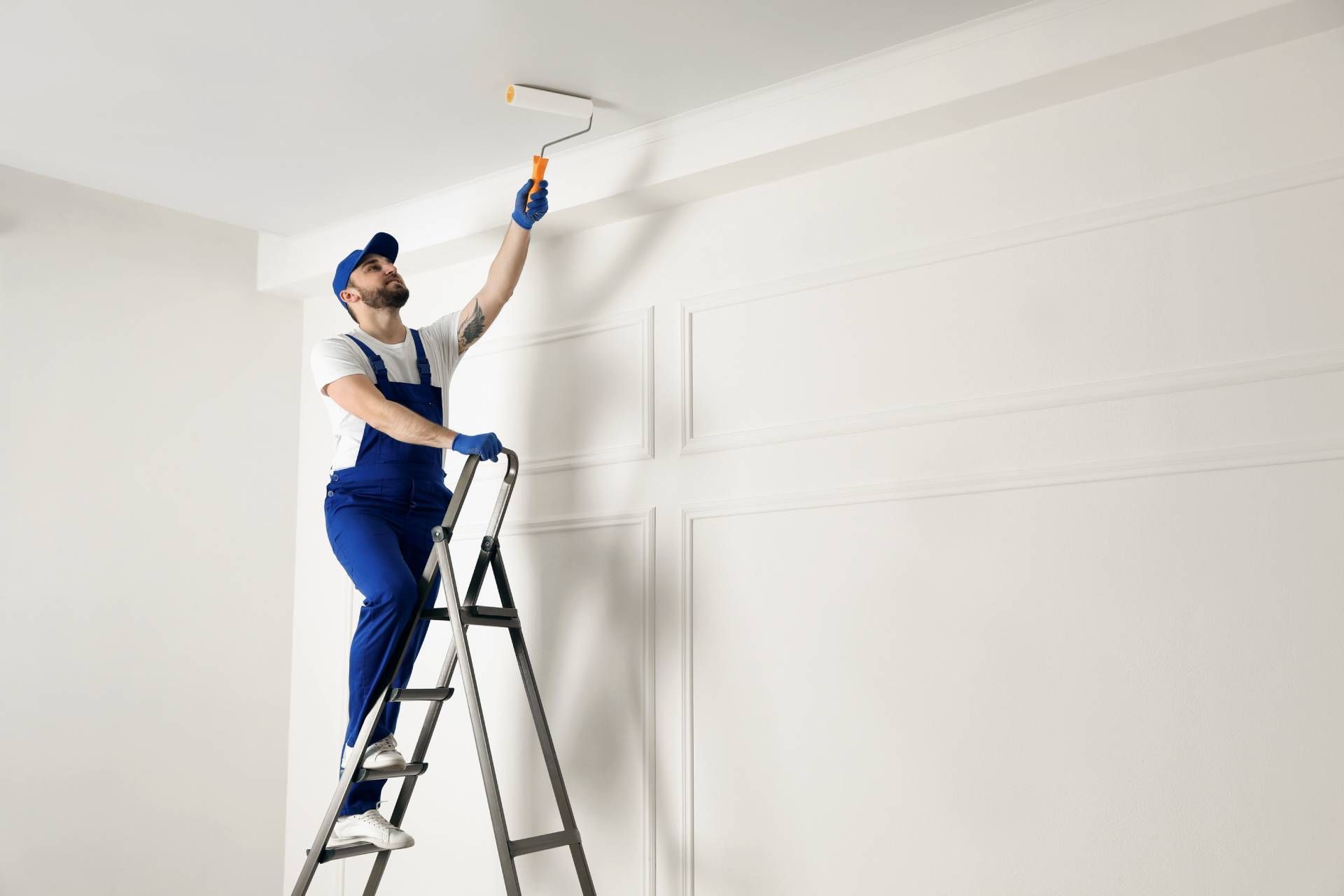 Person on a ladder painting a white ceiling with a roller in a bright room