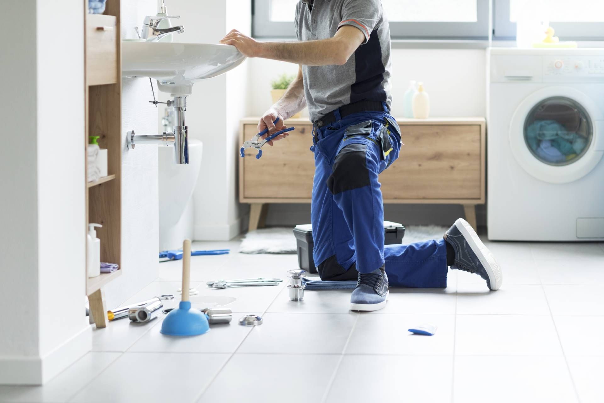 Plumber kneeling under a sink, working on pipes in a bright laundry room with tools on the floor