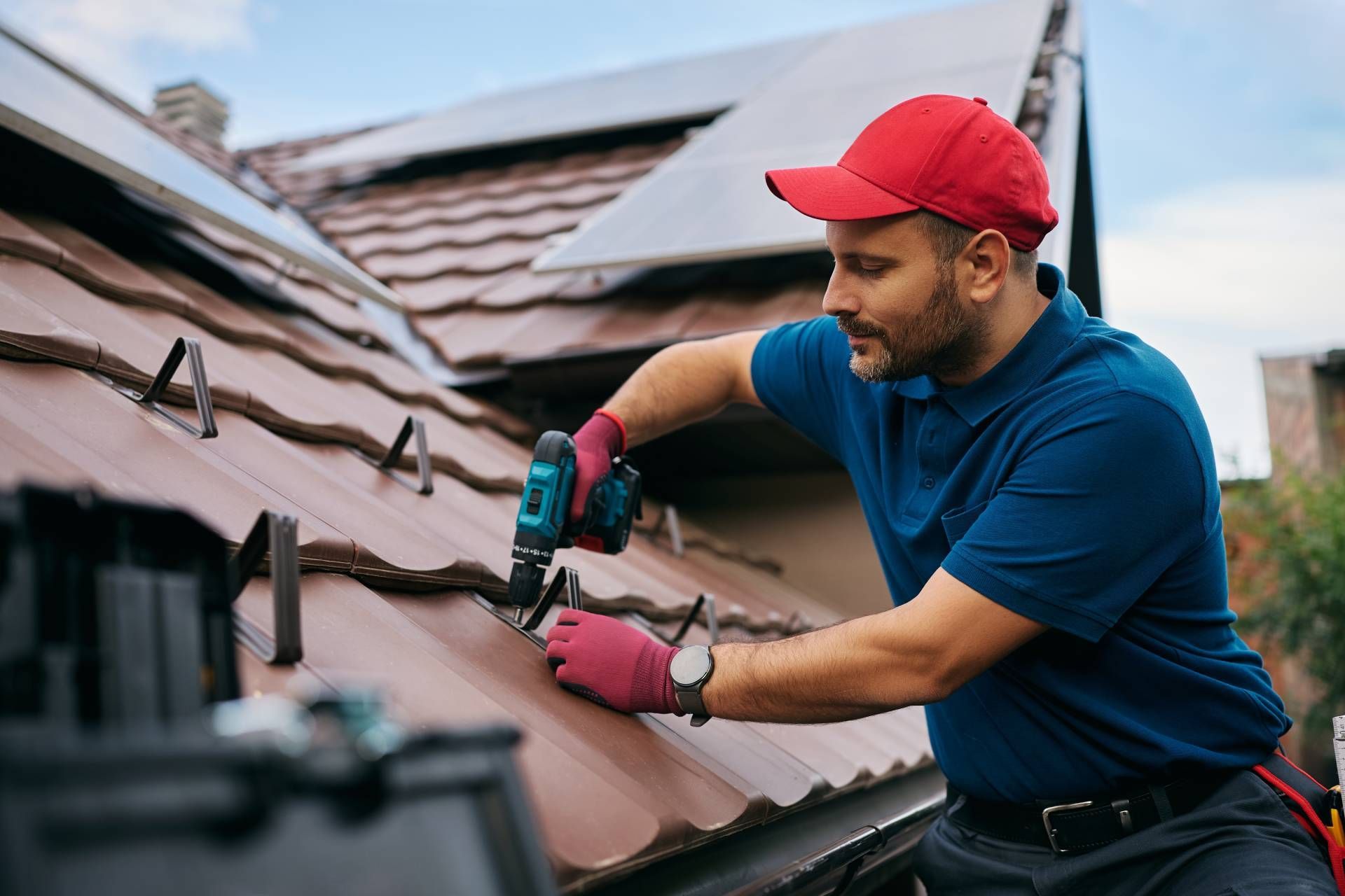 Worker in red cap and blue shirt using a drill to repair roof shingles.