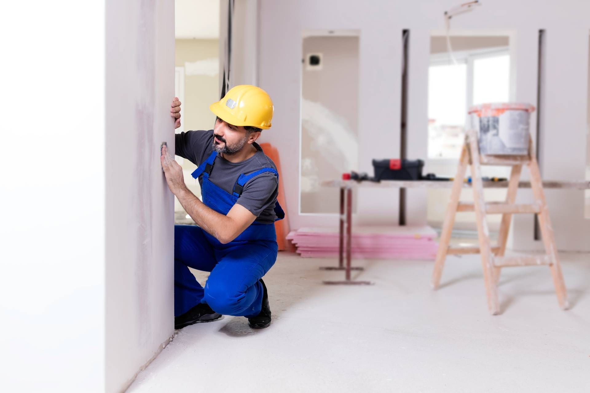 Worker in blue overalls and yellow hard hat measuring a wall in a room under renovation