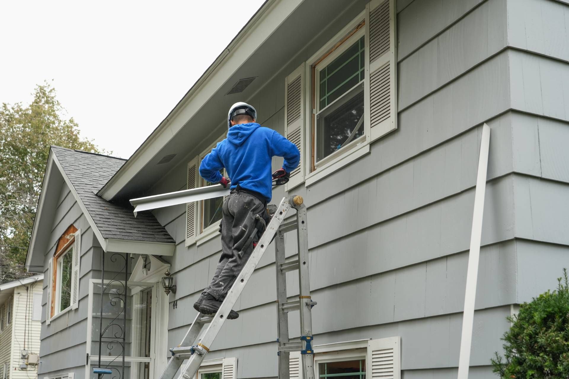 Worker on a ladder painting a gray house exterior near a window and eaves