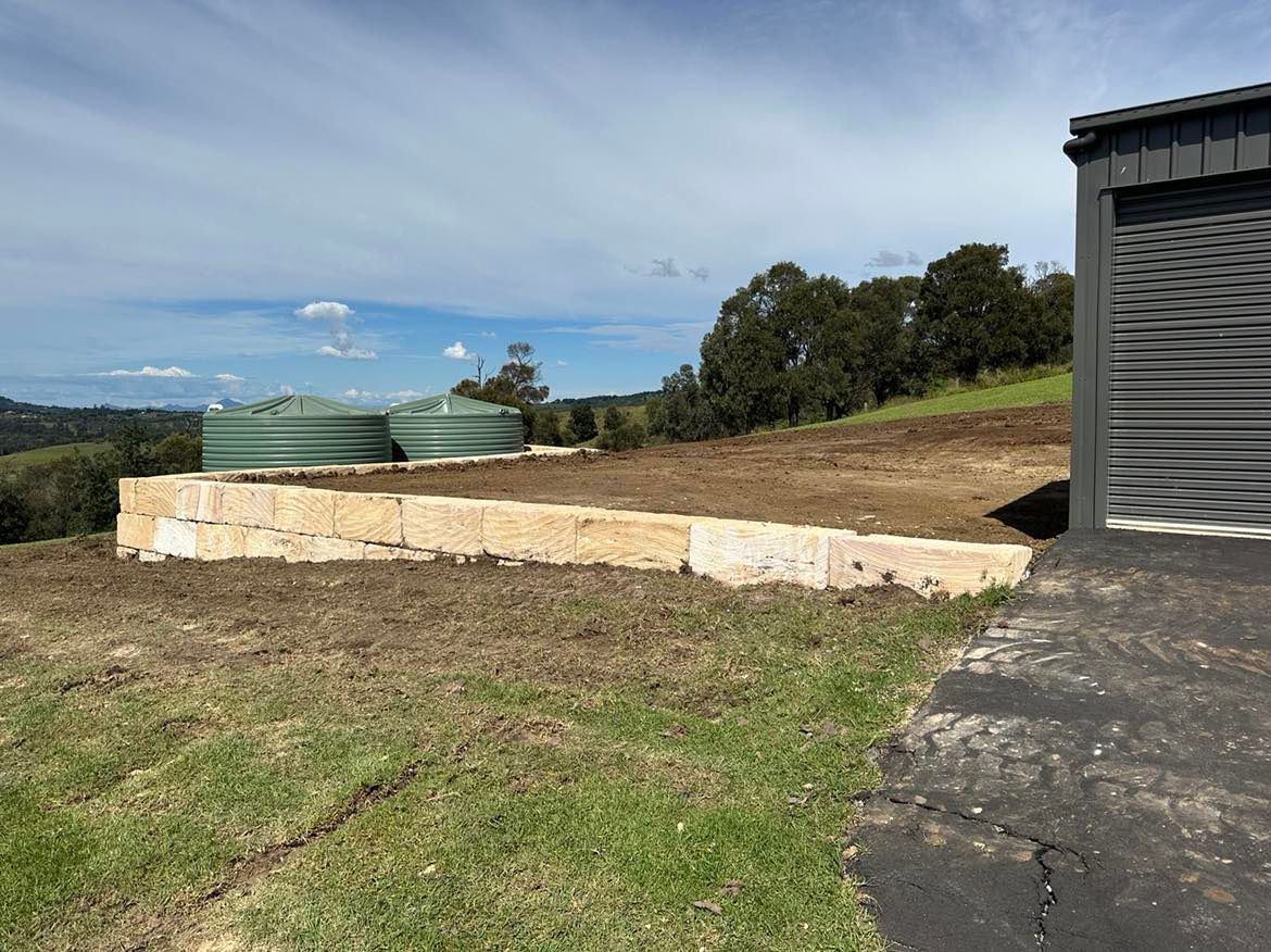 A shed is sitting on top of a dirt hill next to a sandstone wall and a grassy field.