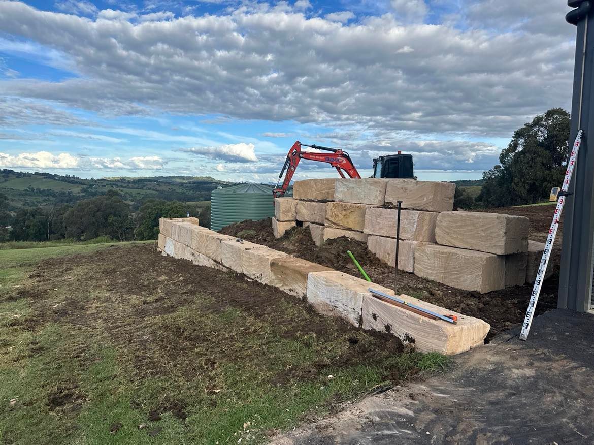 An excavator is working on a sandstone wall in a field.