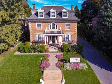 Aerial view of a three-story red brick house with a dark roof and white trim, surrounded by a lawn and purple flowers.