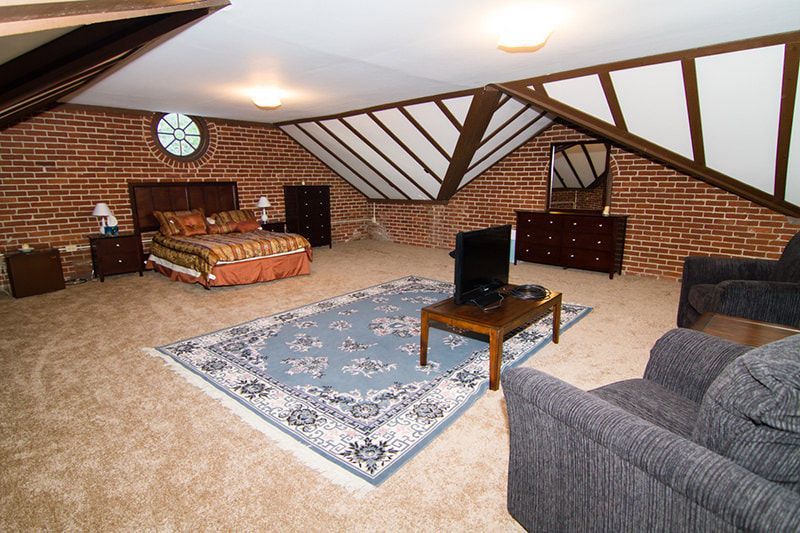 A spacious bedroom in an attic with brick walls, slanted ceilings, a bed, furniture, and a blue rug on carpeted floors.