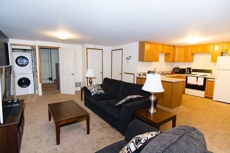 A living room with a dark sofa, coffee table, and lamp, opening into a kitchen with light wood cabinets and a laundry nook.