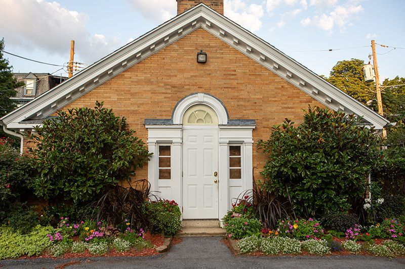 A tan brick building with a central white door, arched transom window, and flanking shrubs under a decorative pediment.