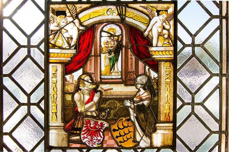 Stained glass showing a knight and a woman kneeling before a central archway with a religious figure inside.
