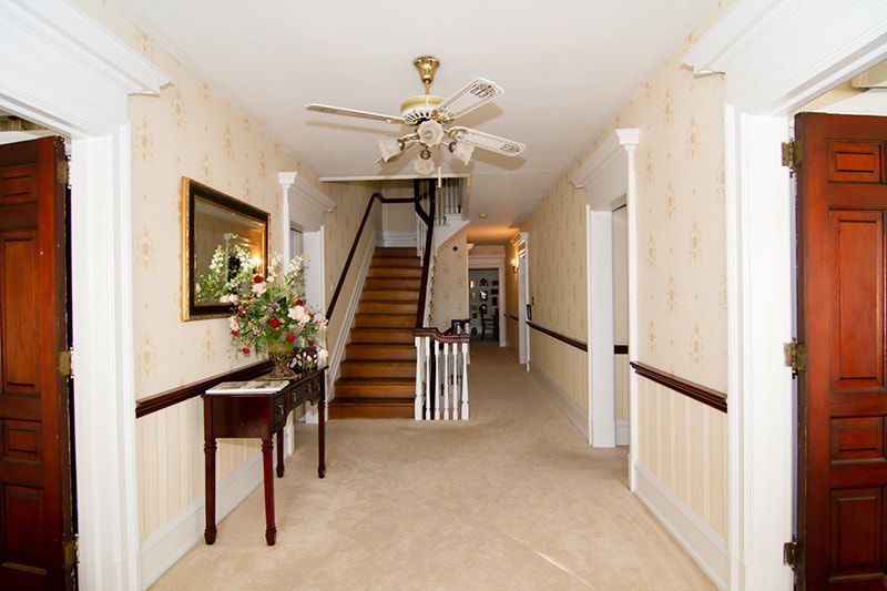 A hallway featuring beige wallpaper, a wooden staircase, a ceiling fan, a console table with flowers, and two wooden doors.