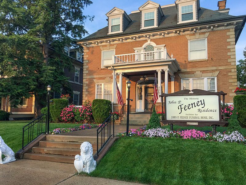 The Feeney Residence, a multi-story brick building with a white porch, American flags, and a front lawn with flowers.