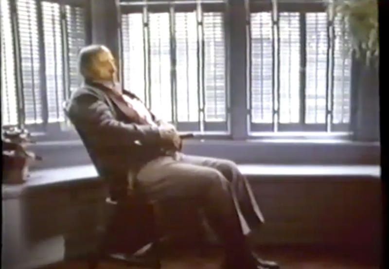 A person sitting in a wooden office chair, facing away from a window with white shutters in a brightly lit room.
