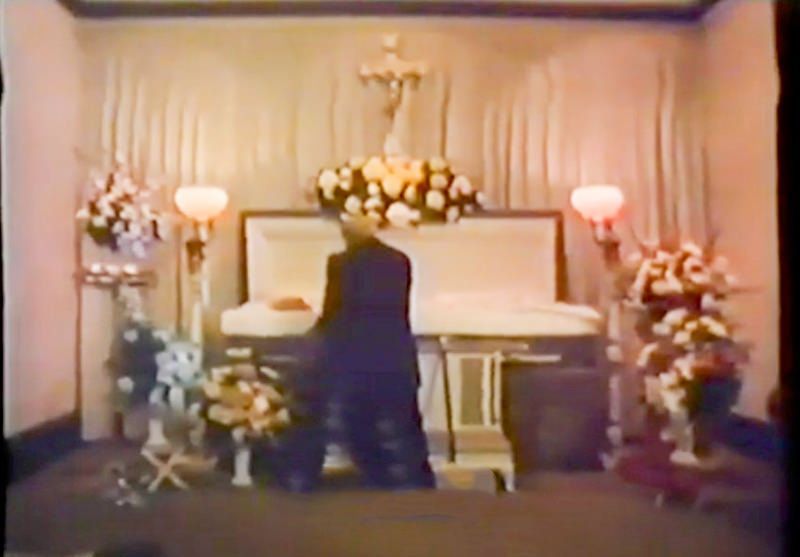 A person in a dark suit kneels before an open casket in a funeral parlor decorated with floral arrangements and a crucifix.