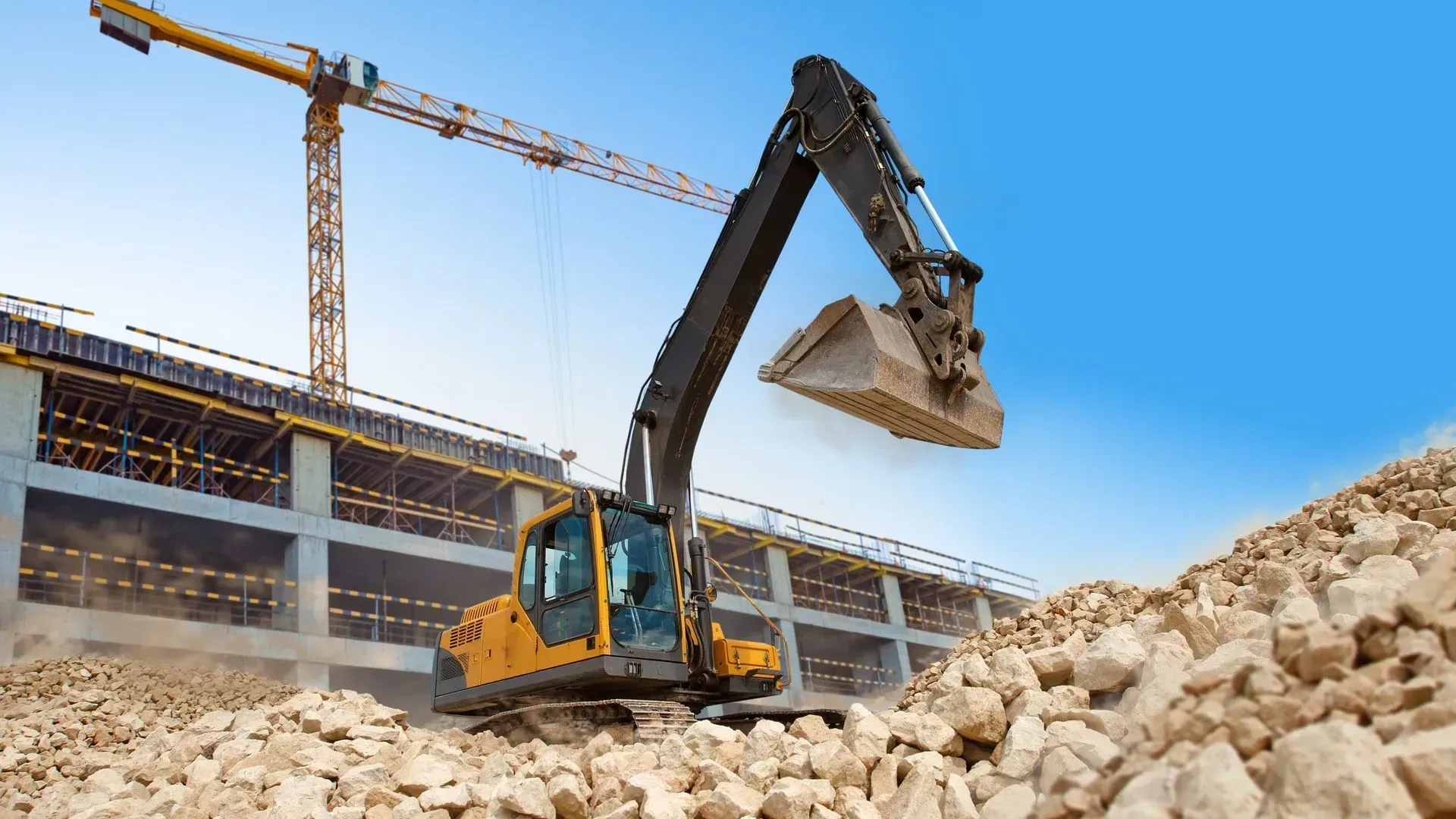 Yellow excavator lifting rubble at a construction site beside a building frame and crane.