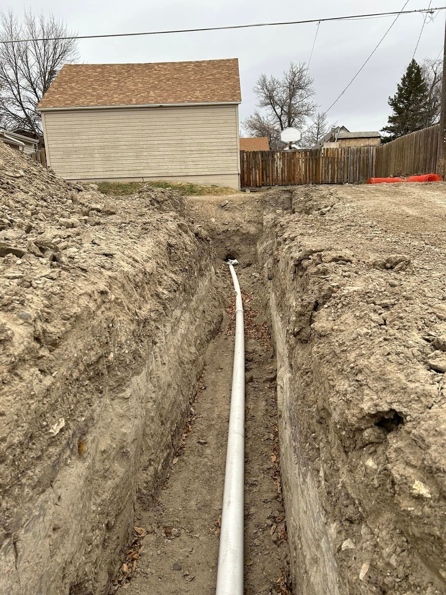 Trench with a white pipe running through dirt toward a beige house and wooden fence in a backyard.