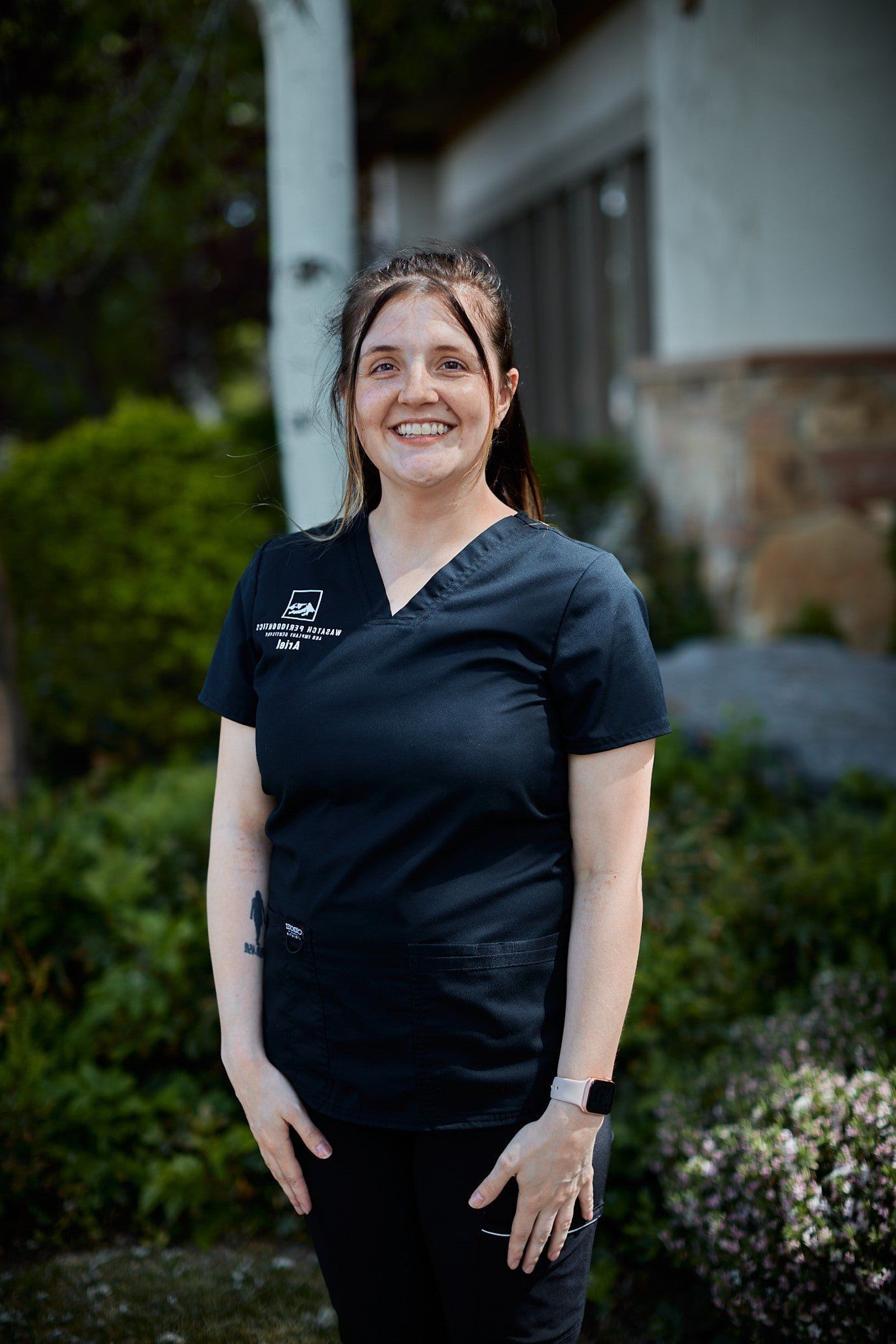 A woman in a black scrub top is smiling and standing in front of a building.
