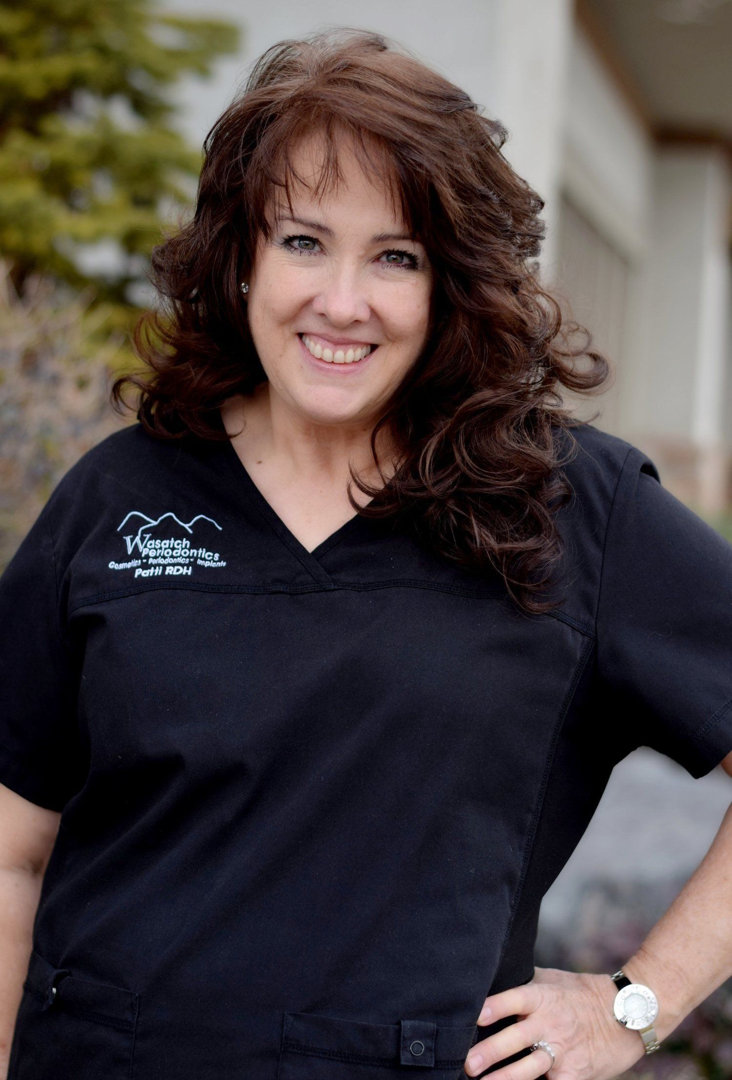 A woman in a black scrub top is smiling and standing with her hands on her hips.