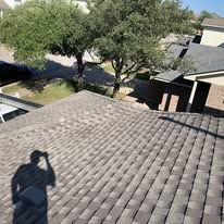A person's shadow on a brown shingled roof with houses and trees in the background.