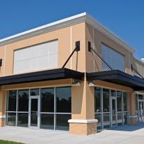 Exterior of a commercial building with beige walls, large windows, and a dark awning under a blue sky.