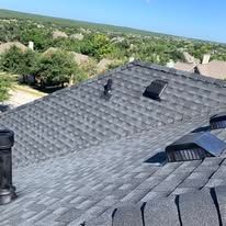 Gray shingle roof with vents and a chimney against a backdrop of green trees and a blue sky.