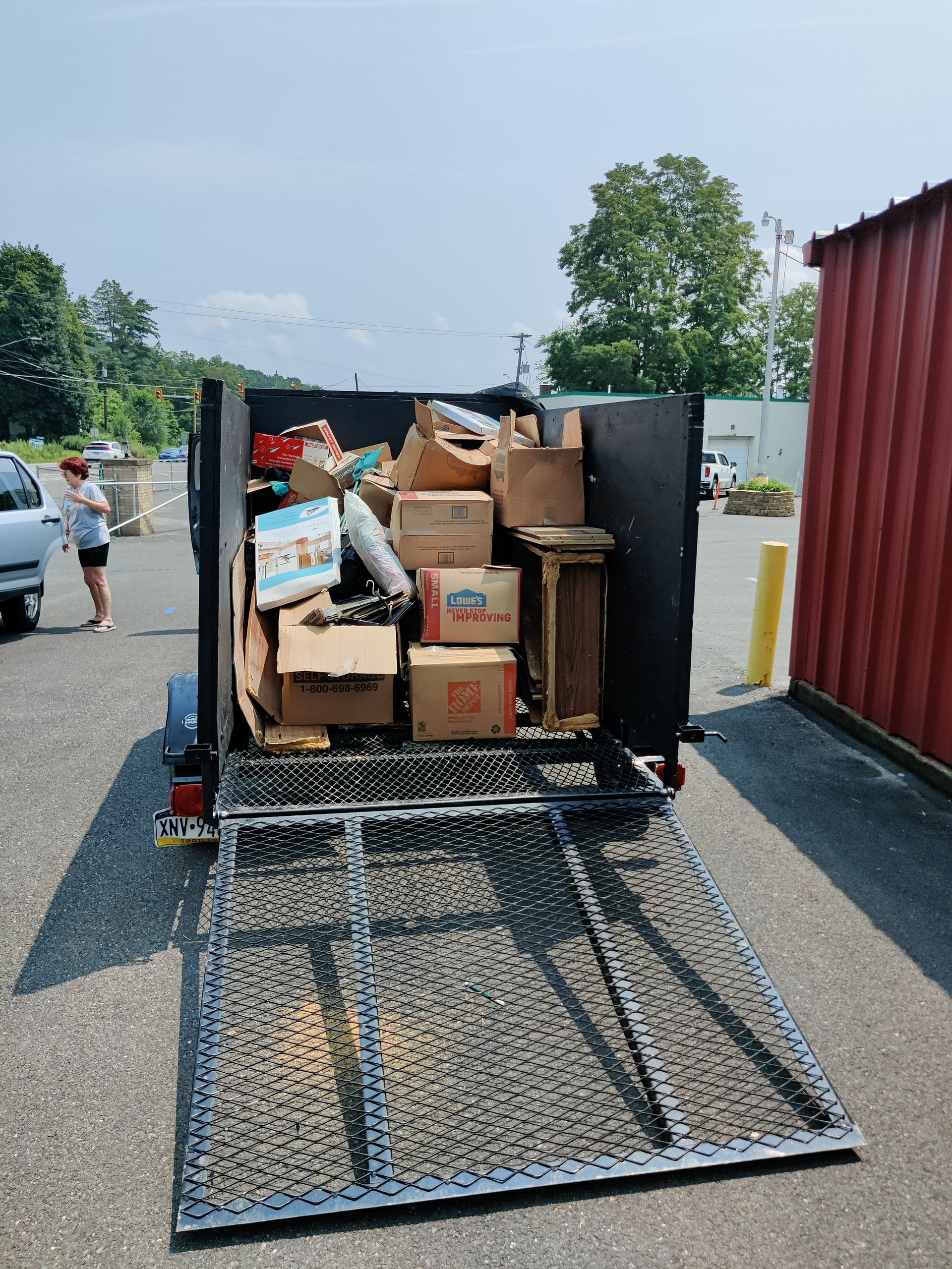 Trailer loaded with boxes and items, ramp down, person nearby, asphalt lot.